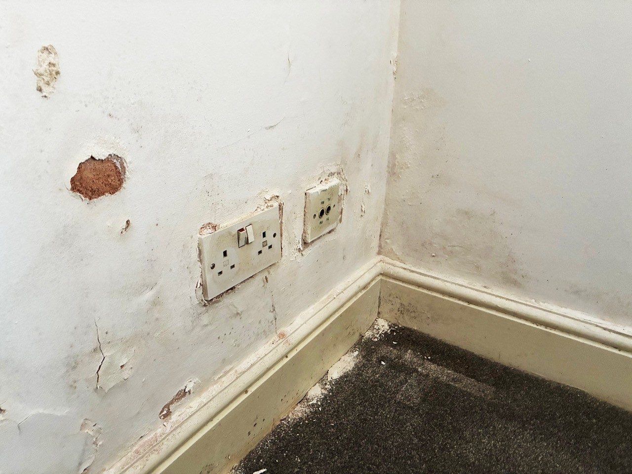 Damp patches and blown plaster on a wall above the skirting, beside two sockets